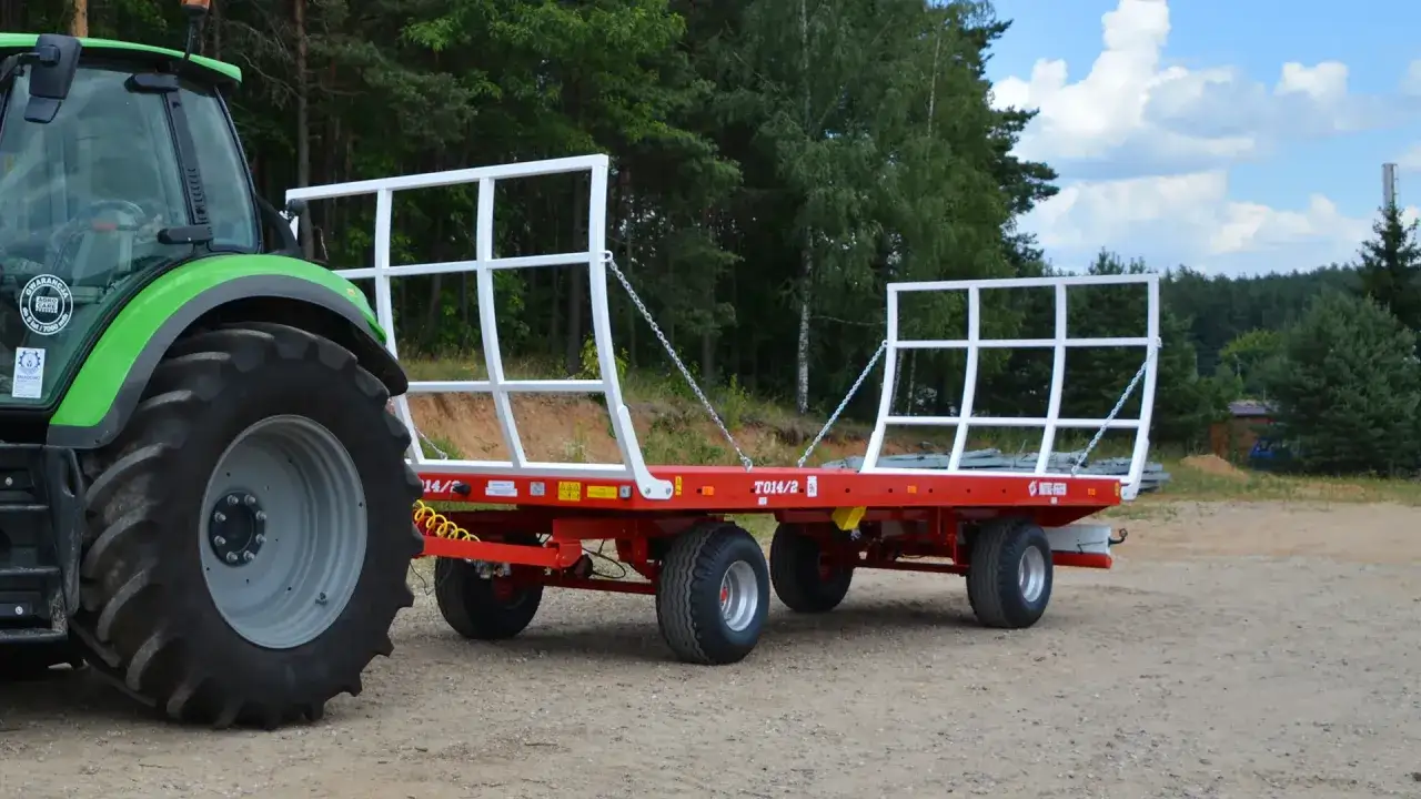 Zielony traktor ciągnie czerwoną przyczepę z białymi burtami. Przyczepa jest pusta, więc nie wiadomo, ile waży balot słomy, który mógłby ją wypełnić.