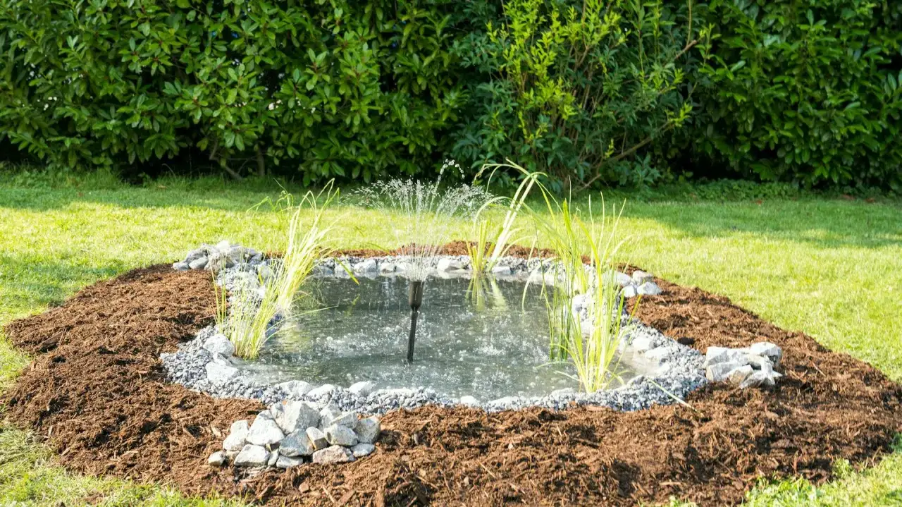 Ein kleiner Gartenbrunnen mit Wasserspiel, umgeben von Rindenmulch und Steinen. Der Teich wurde frisch angelegt und lädt zum Entspannen ein.