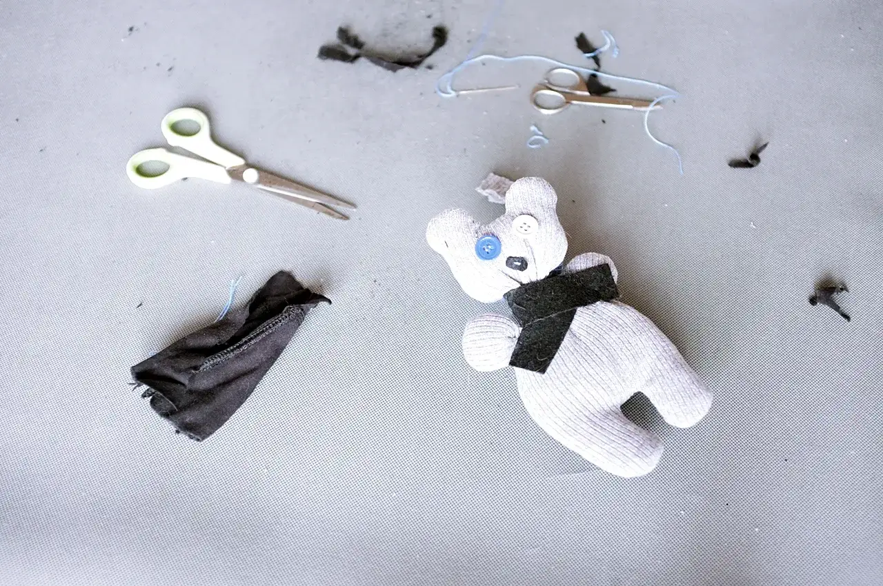 Uroczy pluszak ze skarpety, z guzikami zamiast oczu, leży obok nożyczek i resztek materiału.