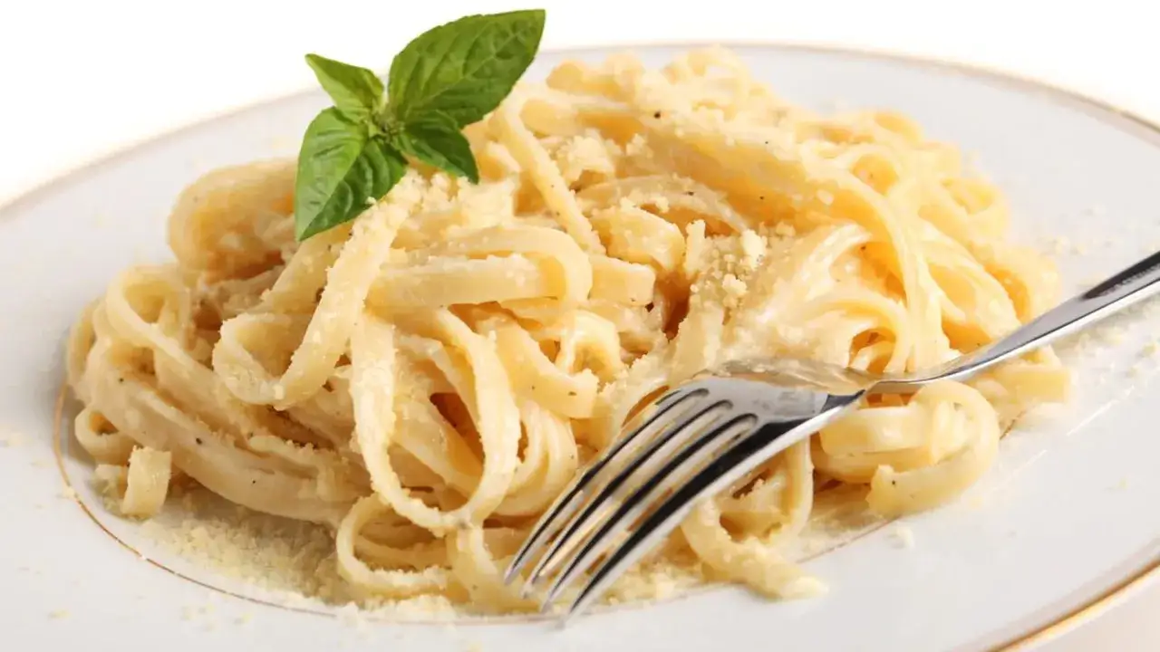 Un piatto di pasta in bianco, condita con formaggio grattugiato e guarnita con una fogliolina di basilico. Una forchetta è appoggiata sul piatto.
