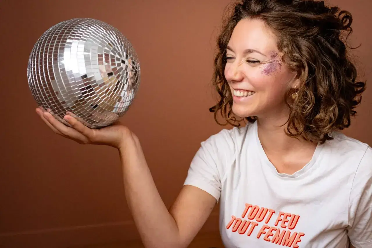Une femme souriante, maquillée de paillettes, tient une boule à facettes. Son t-shirt affiche