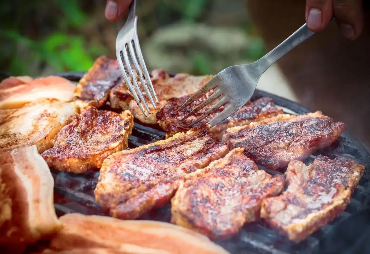 Soczysty boczek na grillu, idealny po marynacie do boczku na grilla. Dwie widelce obracają kawałki mięsa.