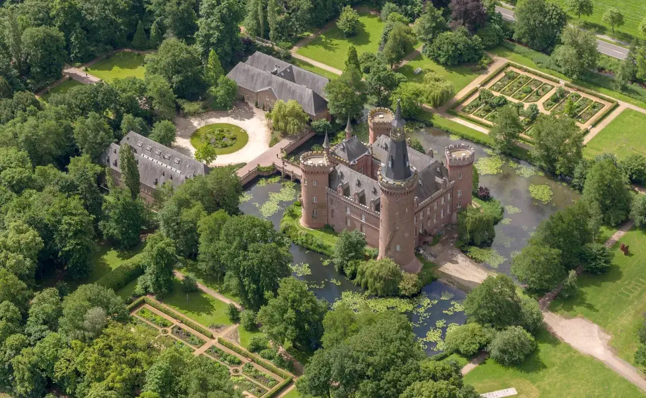 Schloss Moyland, ein märchenhaftes Wasserschloss, umgeben von üppigen Gärten und einem Wassergraben, thront inmitten grüner Bäume.