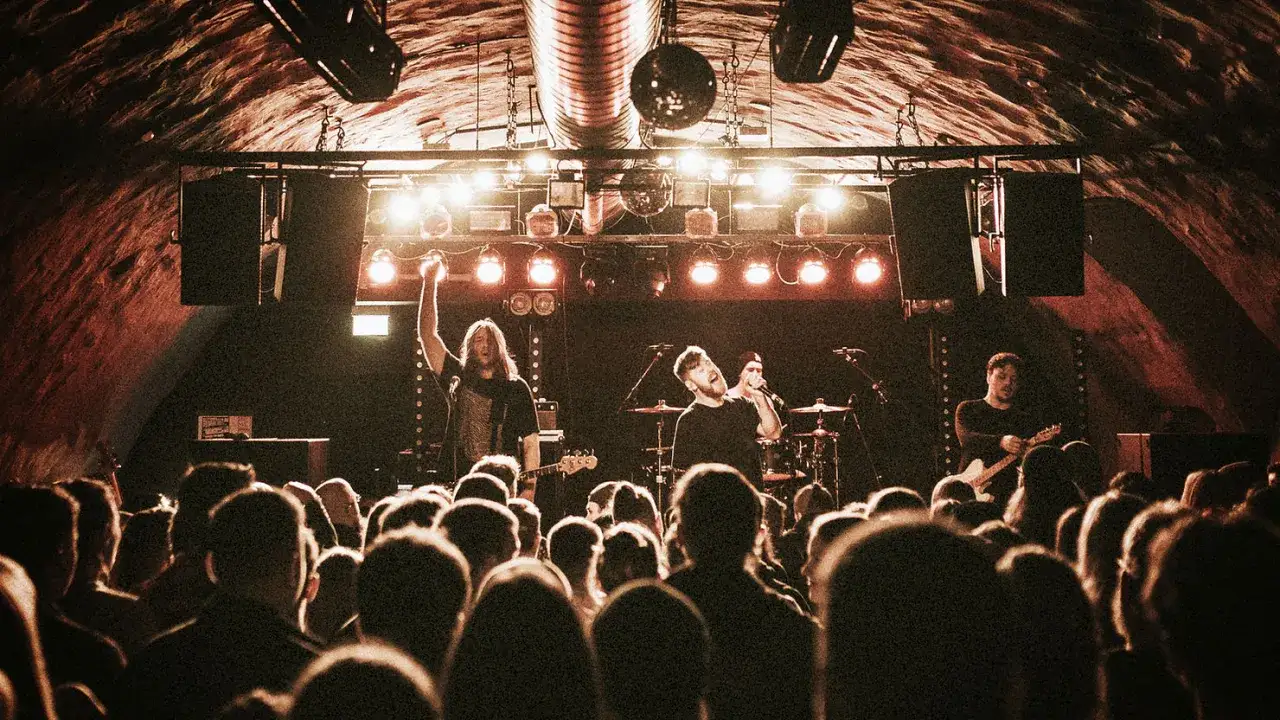 Zespół grający indie rock na koncercie w klubie. Wokalista śpiewa do mikrofonu, gitarzysta gra na gitarze, a perkusista siedzi za zestawem perkusyjnym.