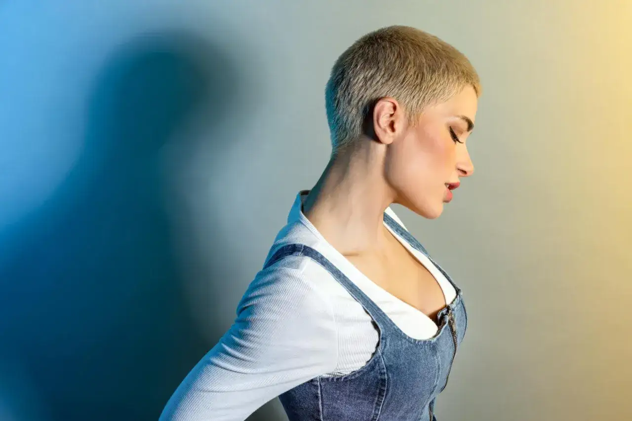 Kobieta z blond buzz cut w dżinsowej sukience na szarym tle z niebieskim i żółtym światłem.