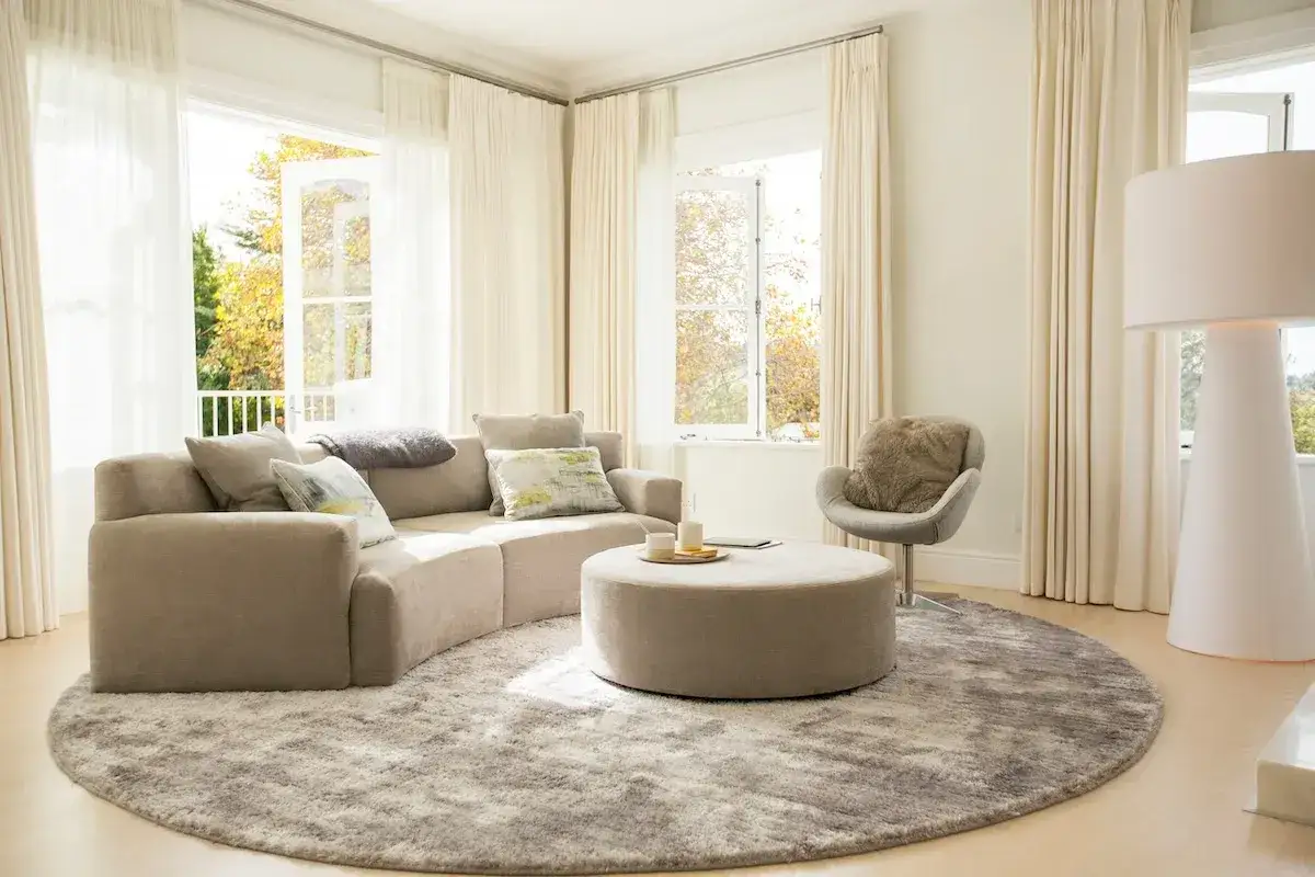 Un soggiorno moderno con divano curvo beige e tappeto rotondo grigio, un esempio perfetto di come abbinare tappeto e divano per creare armonia.