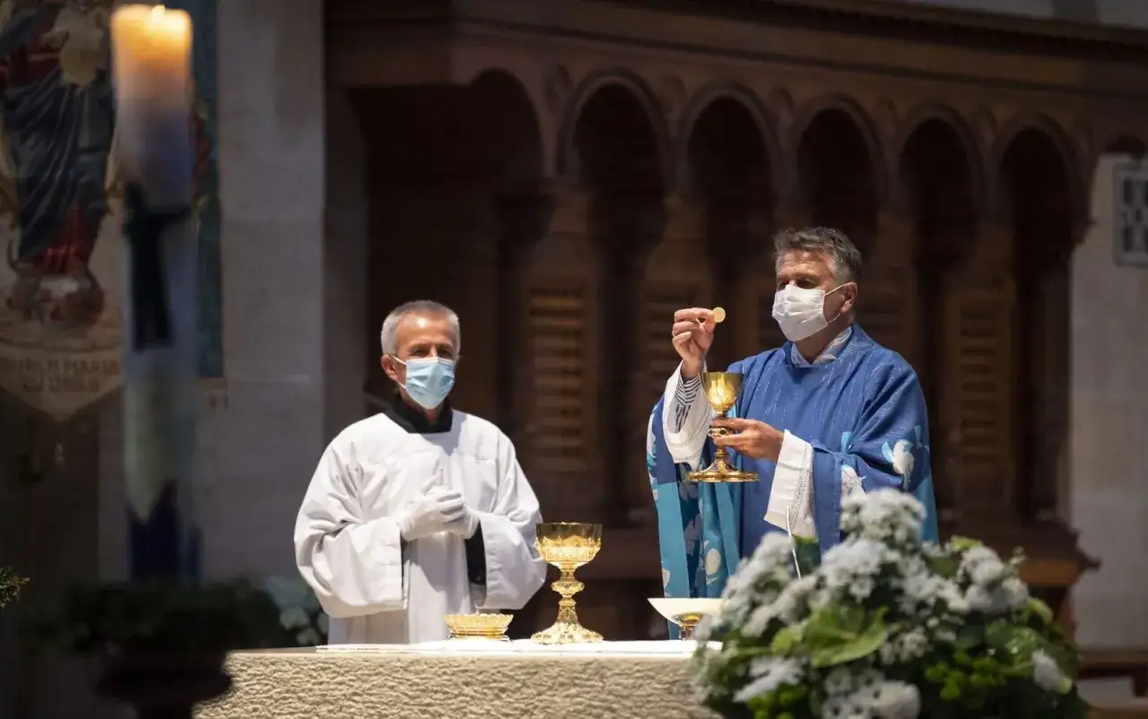 Dwaj księża w maskach podczas mszy. Jeden podnosi hostię do kielicha, celebrując aklamacje po przeistoczeniu.