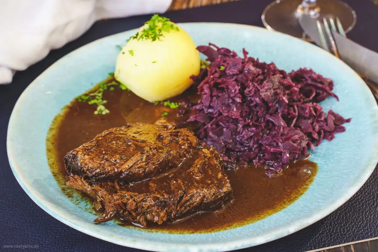 Zarter Rinderbraten in Rotwein eingelegt, serviert mit Klößen und Rotkohl auf einem blauen Teller.