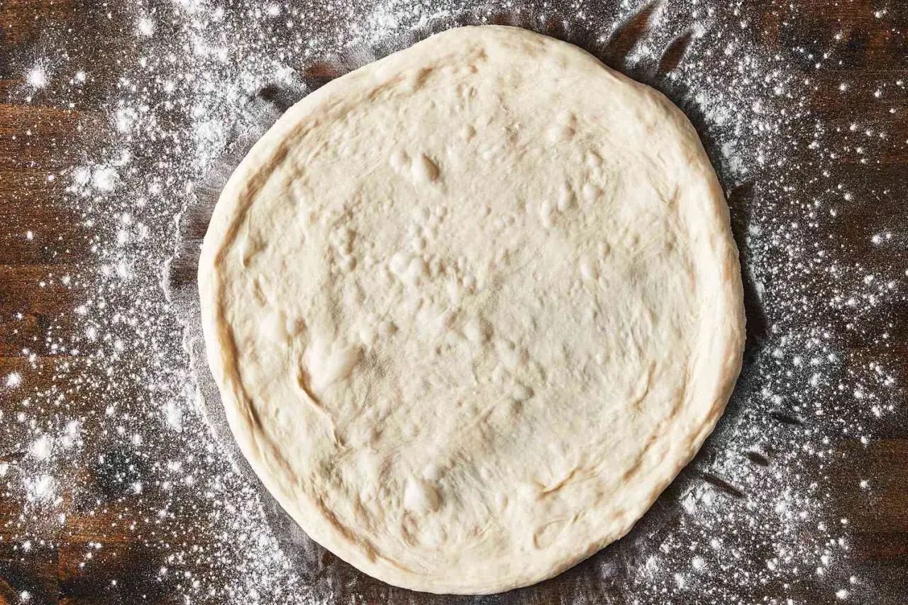Surowe, okrągłe ciasto na pizzę leży na drewnianym blacie, oprószonym mąką. Gotowe na pyszne dodatki!