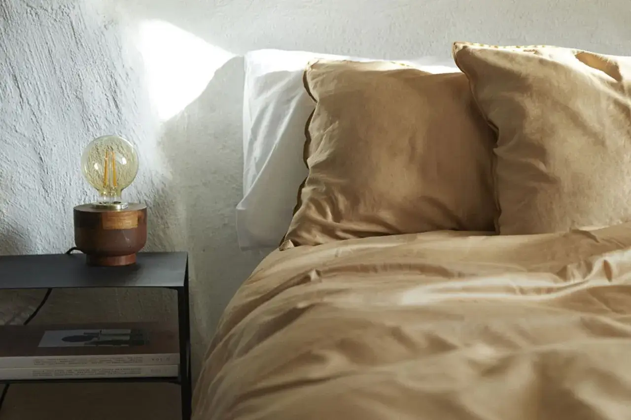 Ein Bett mit ockerfarbener Bettw&auml;sche und Kissen, daneben ein Nachttisch mit Lampe und B&uuml;chern.