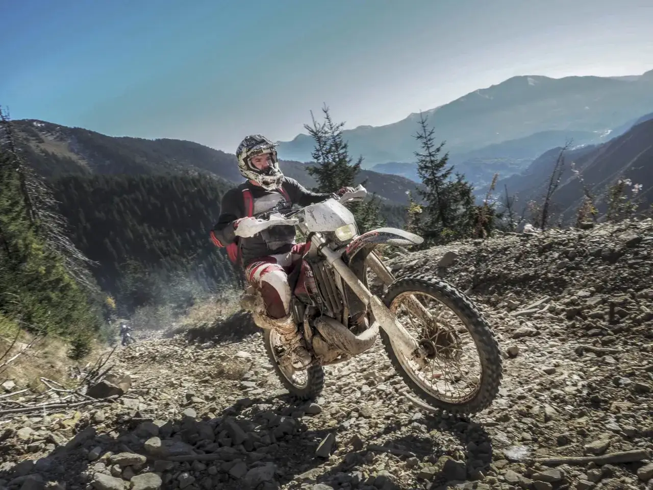 Motocyklista w kasku i ochraniaczach zjeżdża po kamienistym zboczu w górach. Niesamowite widoki i przygoda podczas **camping rumunia**.