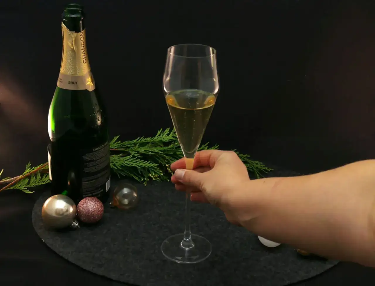 Dłoń trzyma kieliszek musującego **brut champagne** obok butelki i świątecznych ozd&oacute;b.