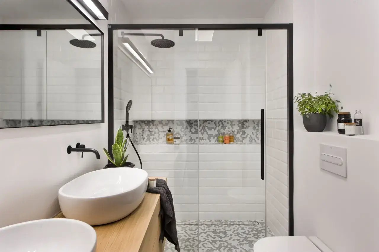 Un elegante cuarto de baño moderno con lavabos sobre encimera de madera, ducha con mampara de cristal y azulejos blancos.