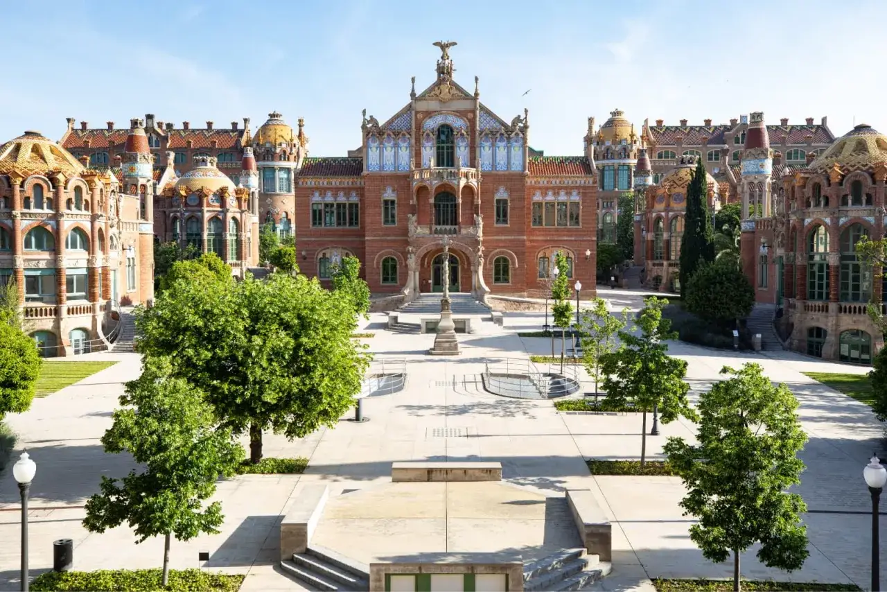 Majestatyczny recinte modernista de Sant Pau z cegły i kolorowych płytek, otoczony zielenią i błękitnym niebem.