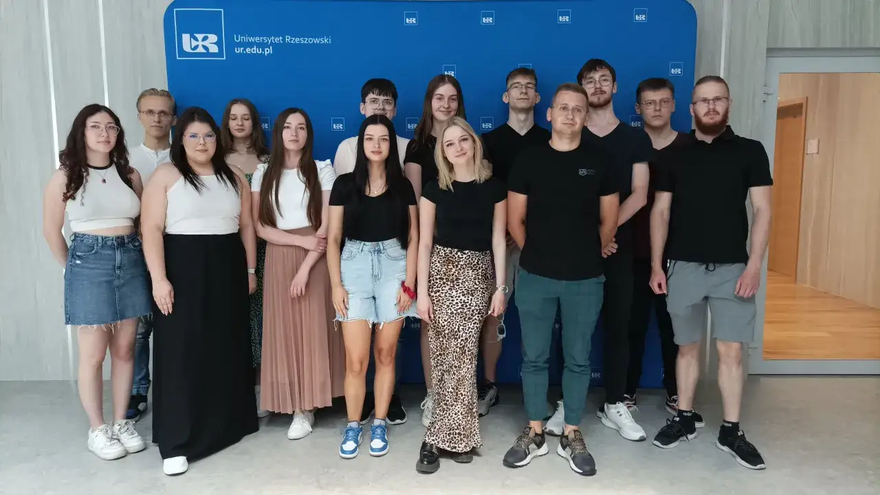 Grupa studentów przed Uniwersytetem Rzeszowskim. Młodzi ludzie pozują do zdjęcia na tle niebieskiej ścianki z logo uczelni.