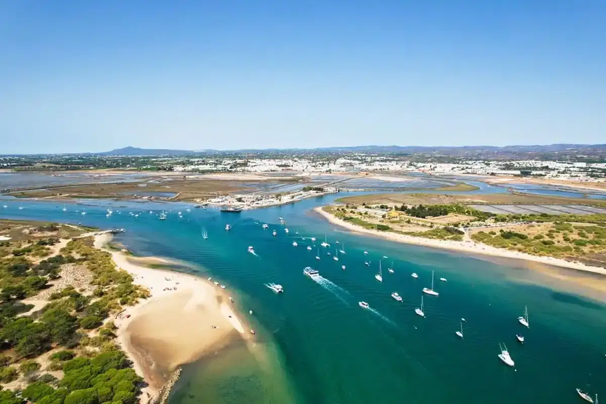 Boote auf dem türkisfarbenen Wasser vor Ilha de Tavira. Eine Sandbank mit grüner Vegetation und die Stadt im Hintergrund.