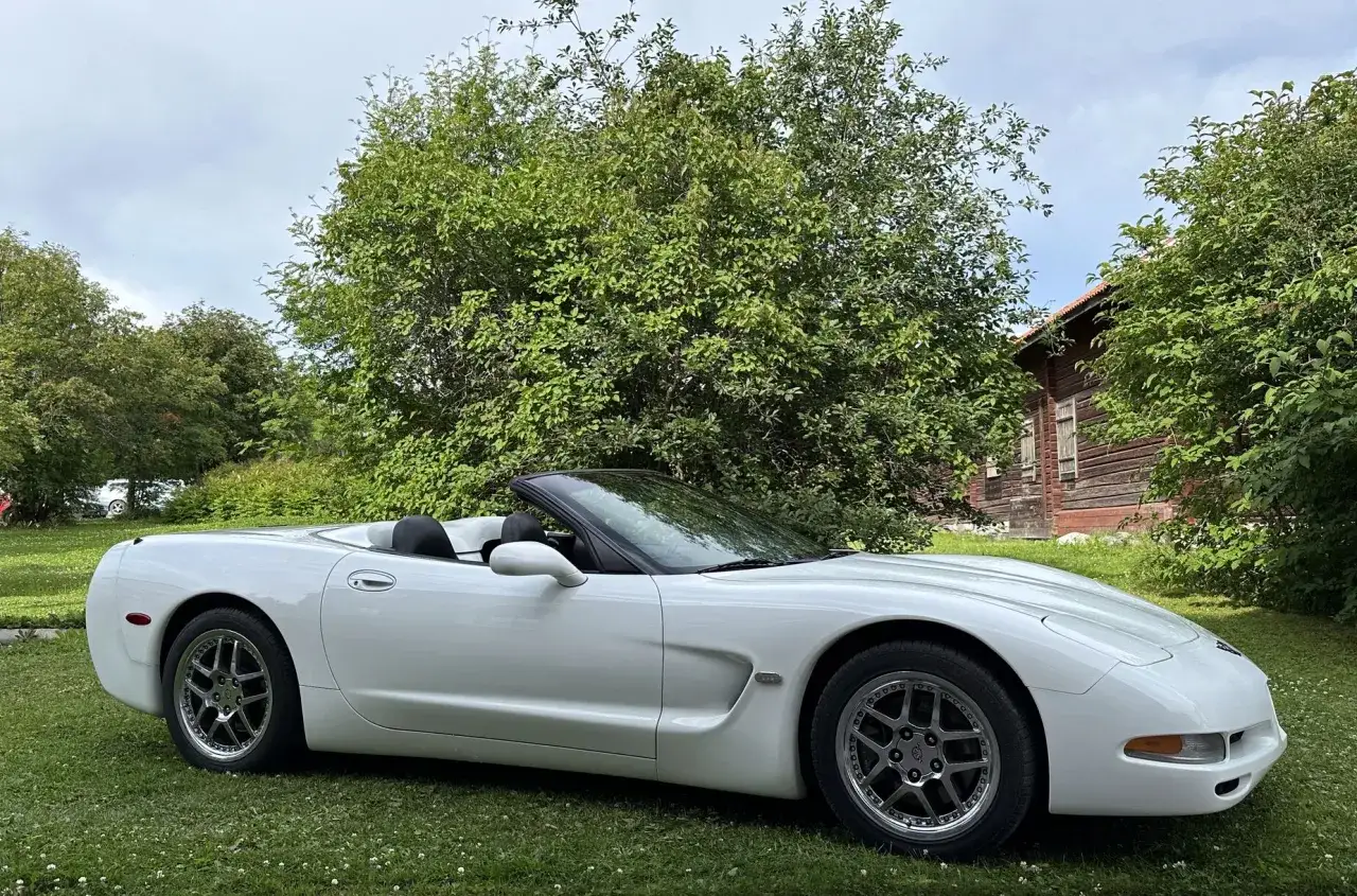 Vit Corvette C5 cabriolet till salu, parkerad p&aring; en gr&auml;smatta framf&ouml;r ett gammalt tr&auml;hus och lummiga tr&auml;d.
