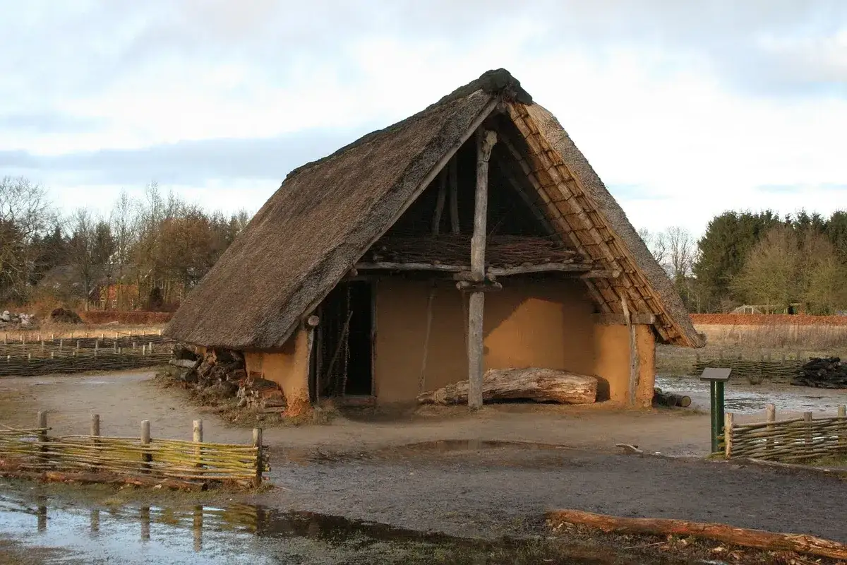 Ein rustikales Bauernhaus aus dem Mittelalter mit Strohdach und Lehmwänden. Holzpfähle stützen das Dach, daneben liegt ein Baumstamm.