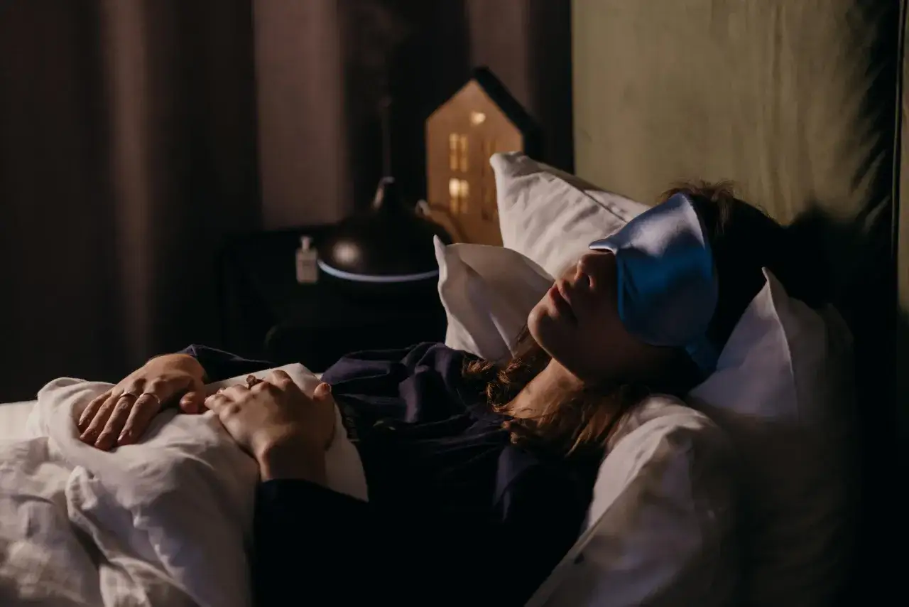 Una mujer duerme con antifaz, soñando con una persona fallecida y verla viva. A su lado, un difusor de aromas y una casa de luz.