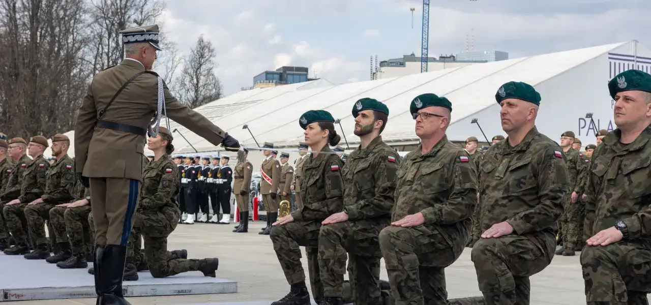 Uroczystość wojskowa, gdzie żołnierze klęczą przed oficerem. Jeden z nich, w mundurze, symbolizuje najniższy stopień oficerski.