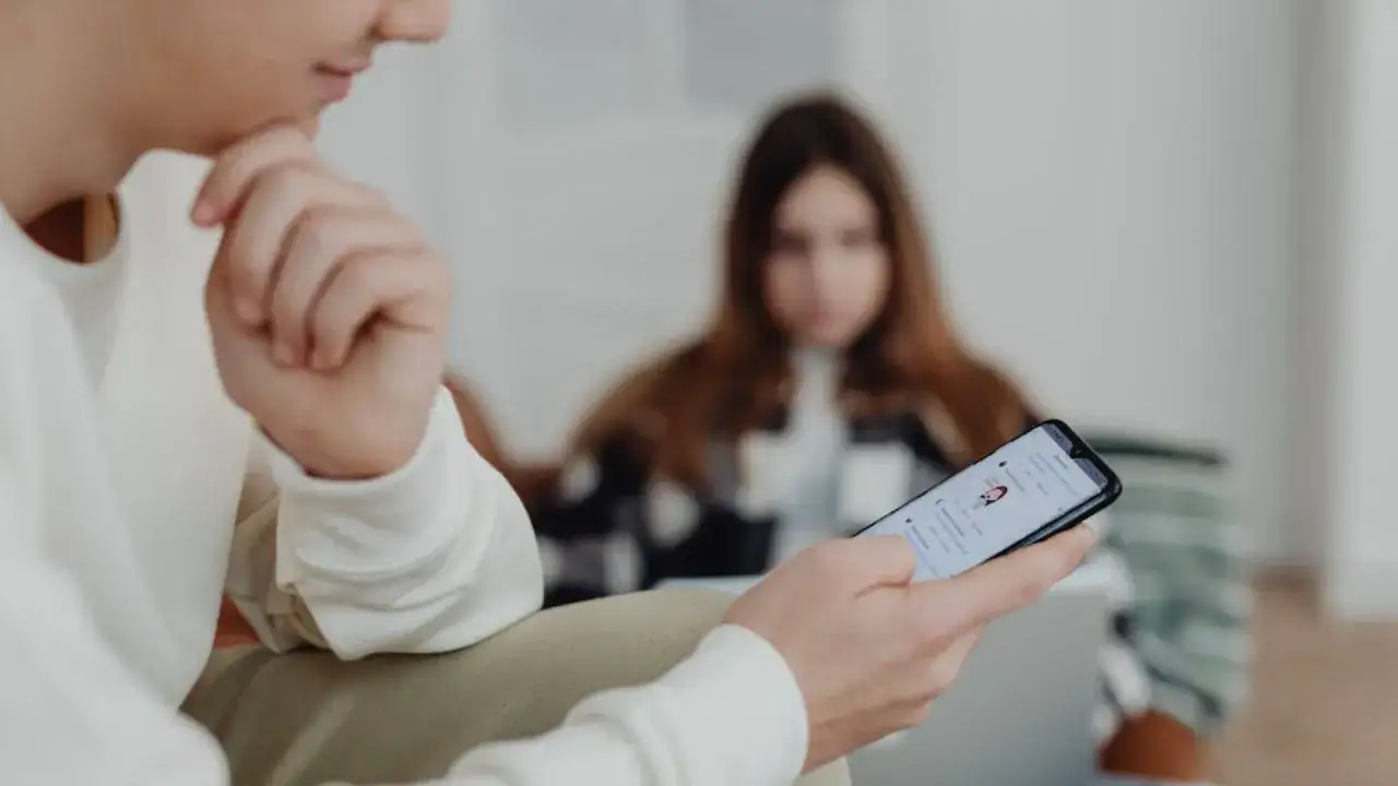 Un jeune homme r&eacute;fl&eacute;chit, son t&eacute;l&eacute;phone affichant une liste de contacts, illustrant la **hyperconnectivit&eacute; d&eacute;finition**.