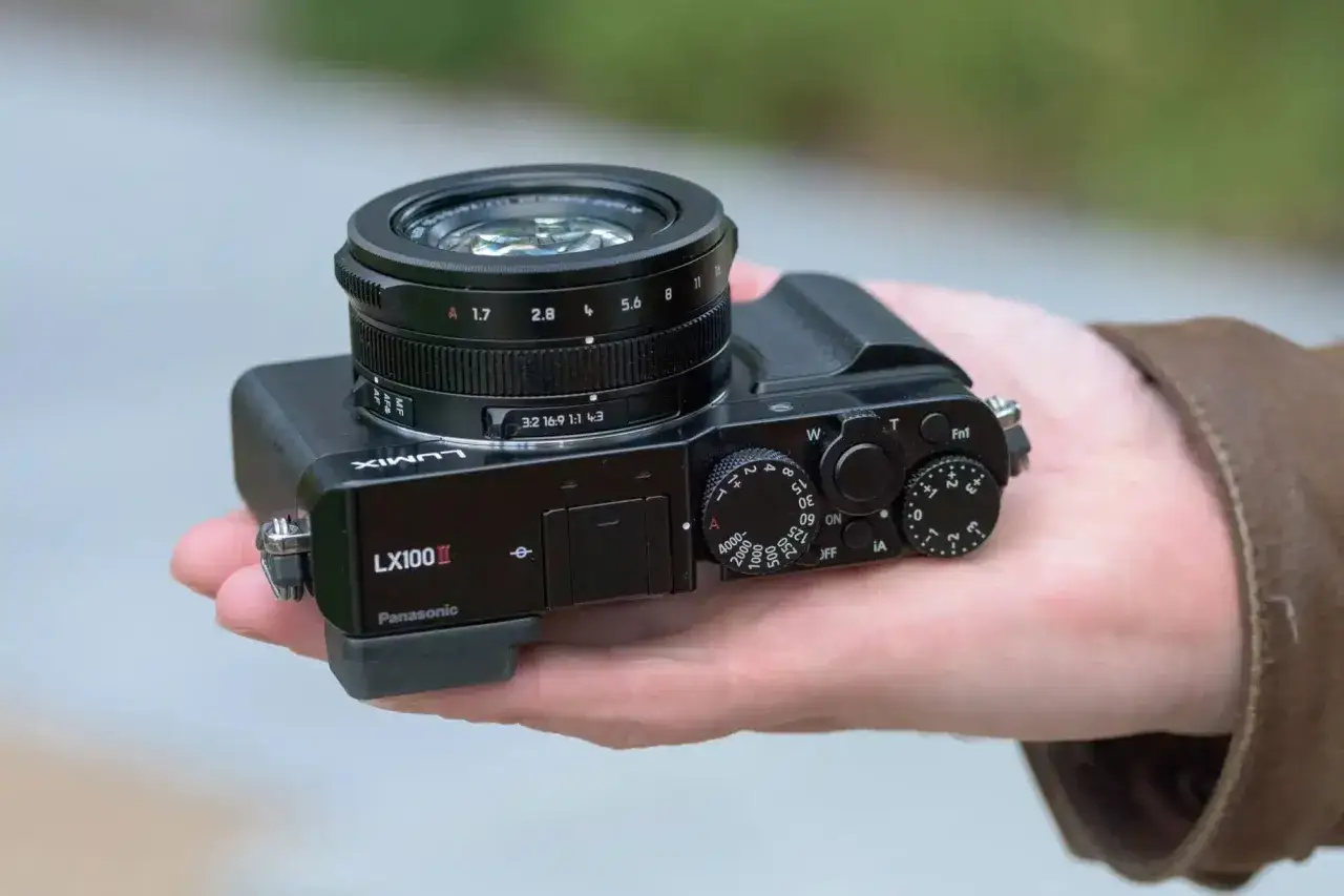 A hand holds a black Panasonic Lumix DMC-LX100K camera, showcasing its lens and top controls.