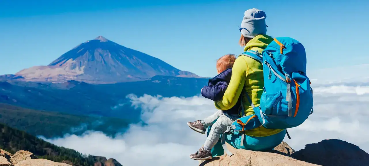 Madre con bebé en mochila, disfrutando del turismo activo con vistas a un volcán y nubes.