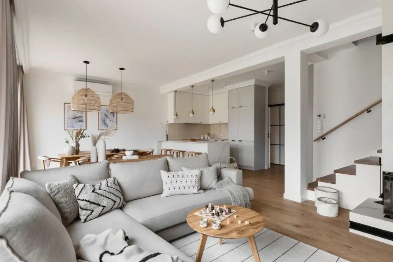 Przytulny, **soft loft** z szarą kanapą, drewnianym stolikiem i otwartą kuchnią. Na stole szachy, w tle schody.