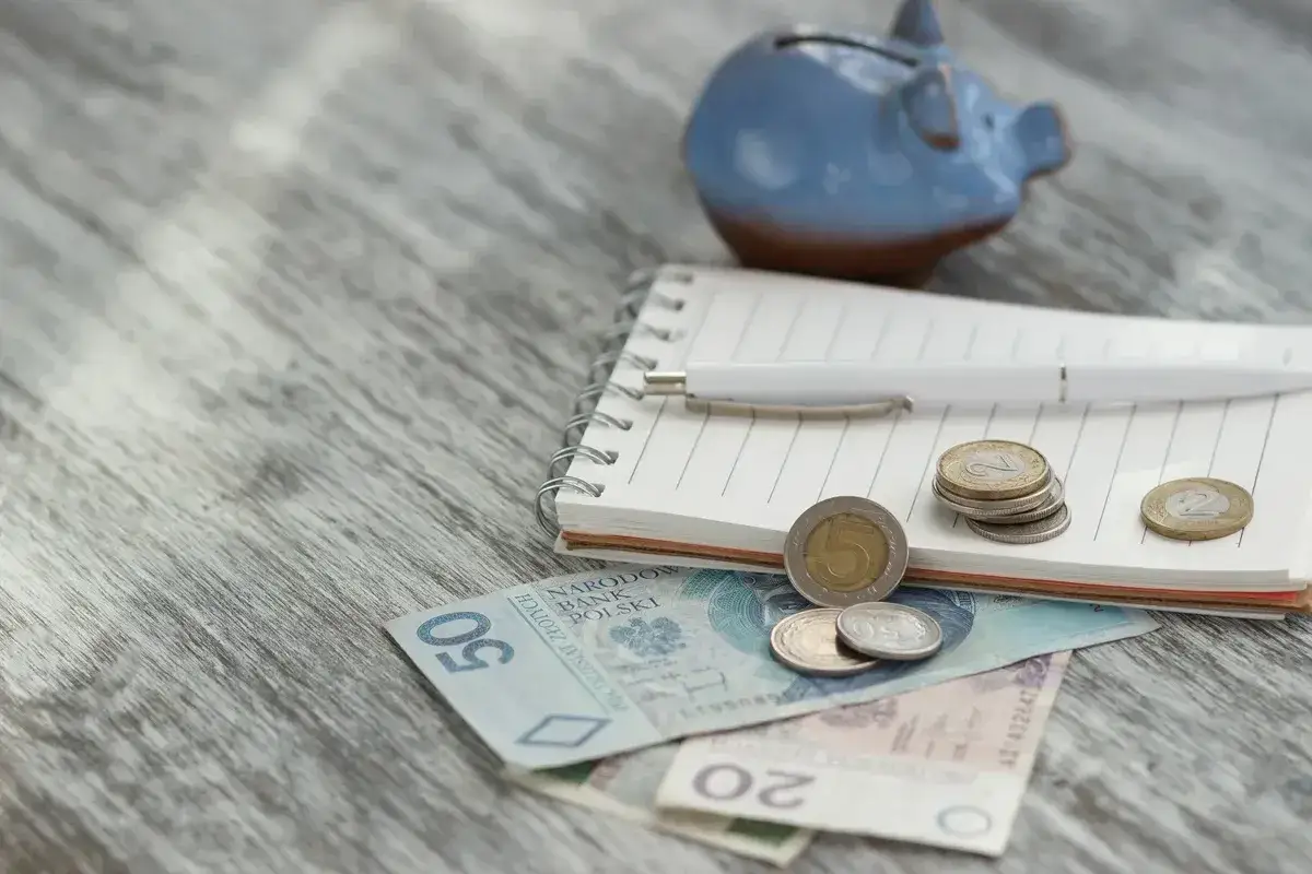 Zapiski o rentę socjalną z MOPS. Na drewnianym blacie leżą polskie banknoty, monety, notatnik i niebieskowa świnka-skarbonka.