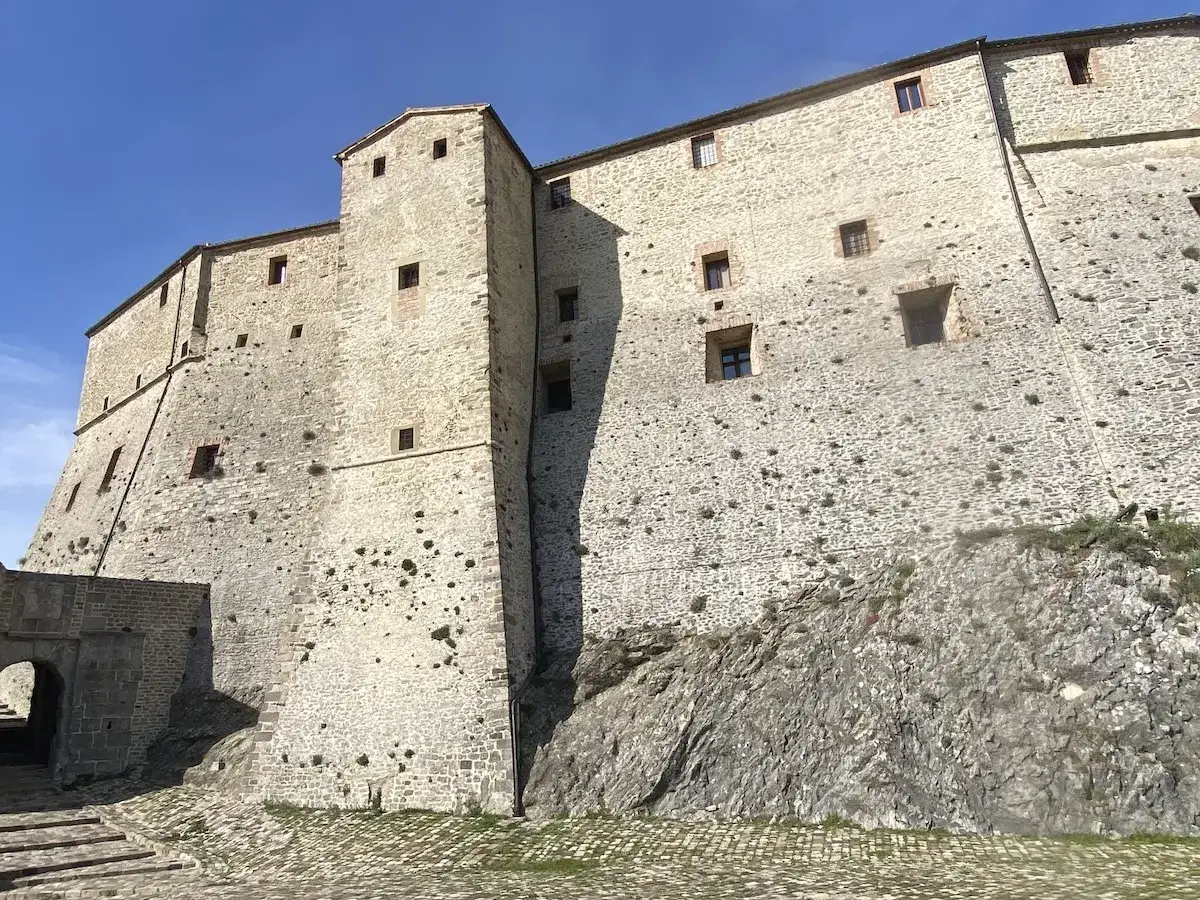 Imponente rocca di **Borgo San Leo** su uno sperone roccioso, con mura in pietra chiara e piccole finestre quadrate sotto un cielo azzurro.