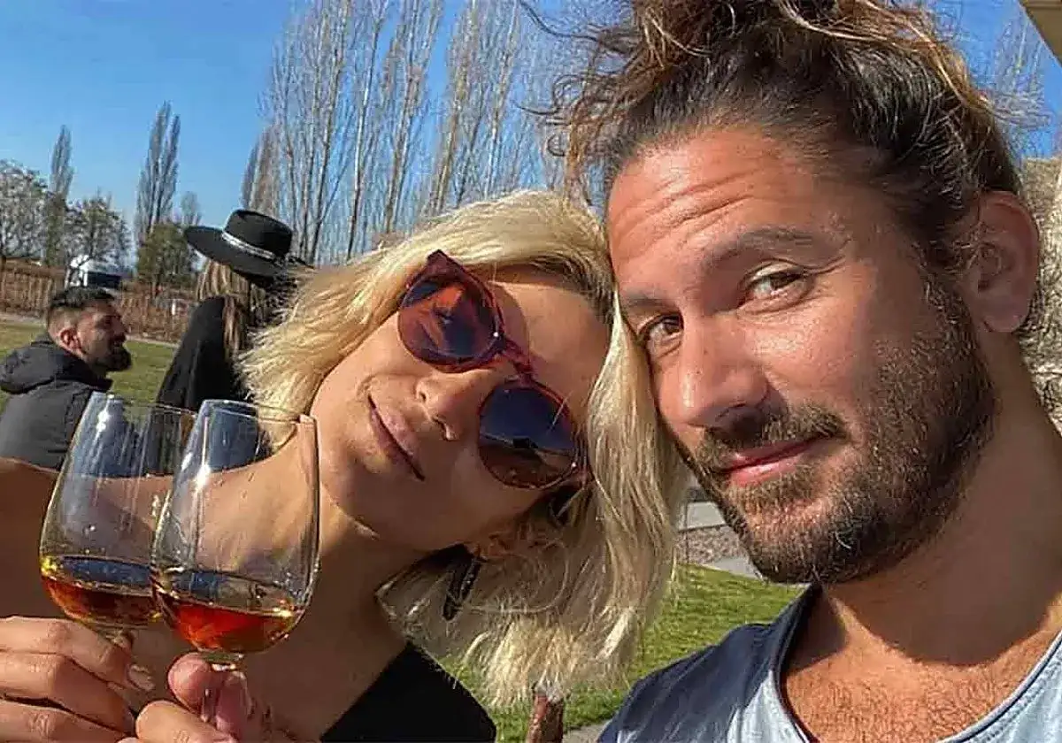 Pareja feliz en un d&iacute;a soleado, brindando con copas de vino. El lago y los &aacute;rboles crean un fondo id&iacute;lico.