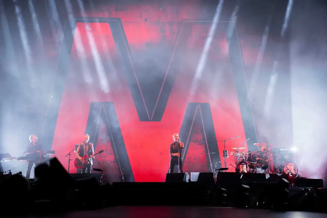 Scenljus lyser upp bandet under en Depeche Mode konsert. En stor r&ouml;d M-logotyp syns bakom dem.