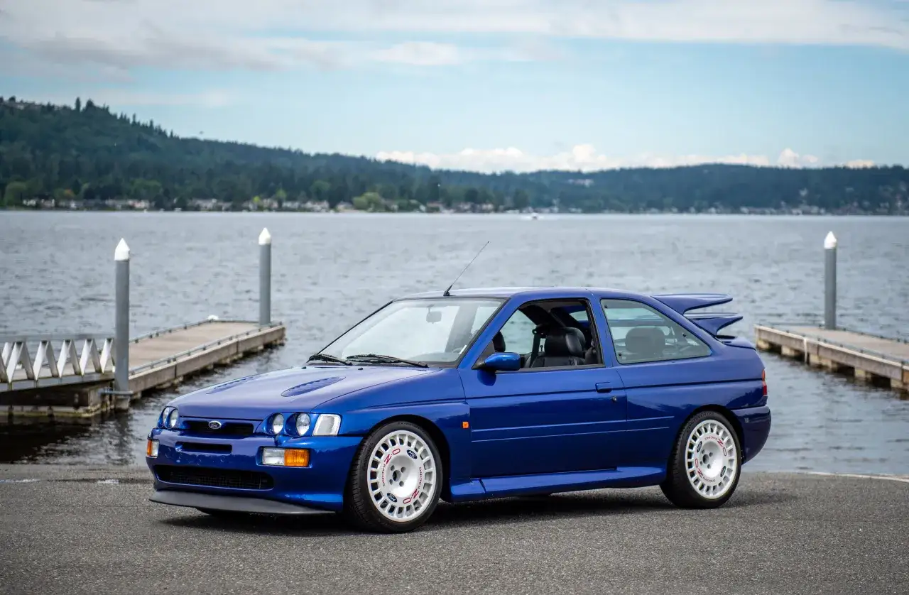 Blå Ford Escort Cosworth parkerad vid en brygga med en sjö och skog i bakgrunden.