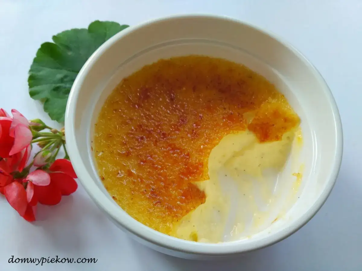 Chrupiąca skorupka karmelizowanego cukru na kremowym creme brulee, obok czerwonych kwiatów i zielonego liścia.
