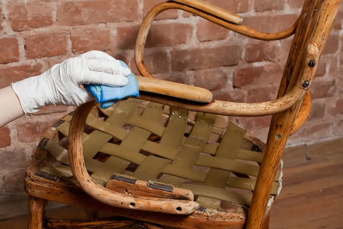 Mano con guante blanco limpia un sillón de madera antiguo, preparándolo para decapar madera y restaurar su belleza original.