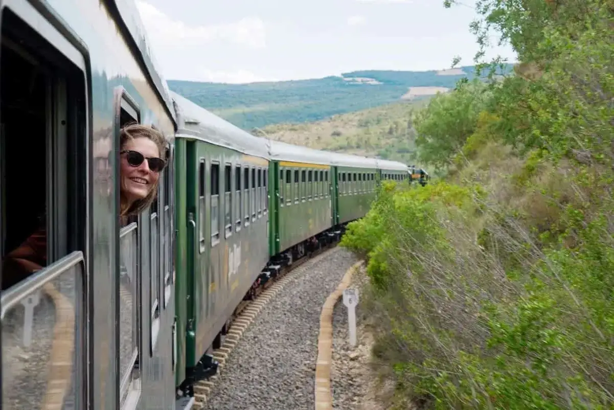 A woman smiles from a train window, enjoying the scenery on weekend trips from Barcelona by train. The green train winds through a lush, hilly landscape.