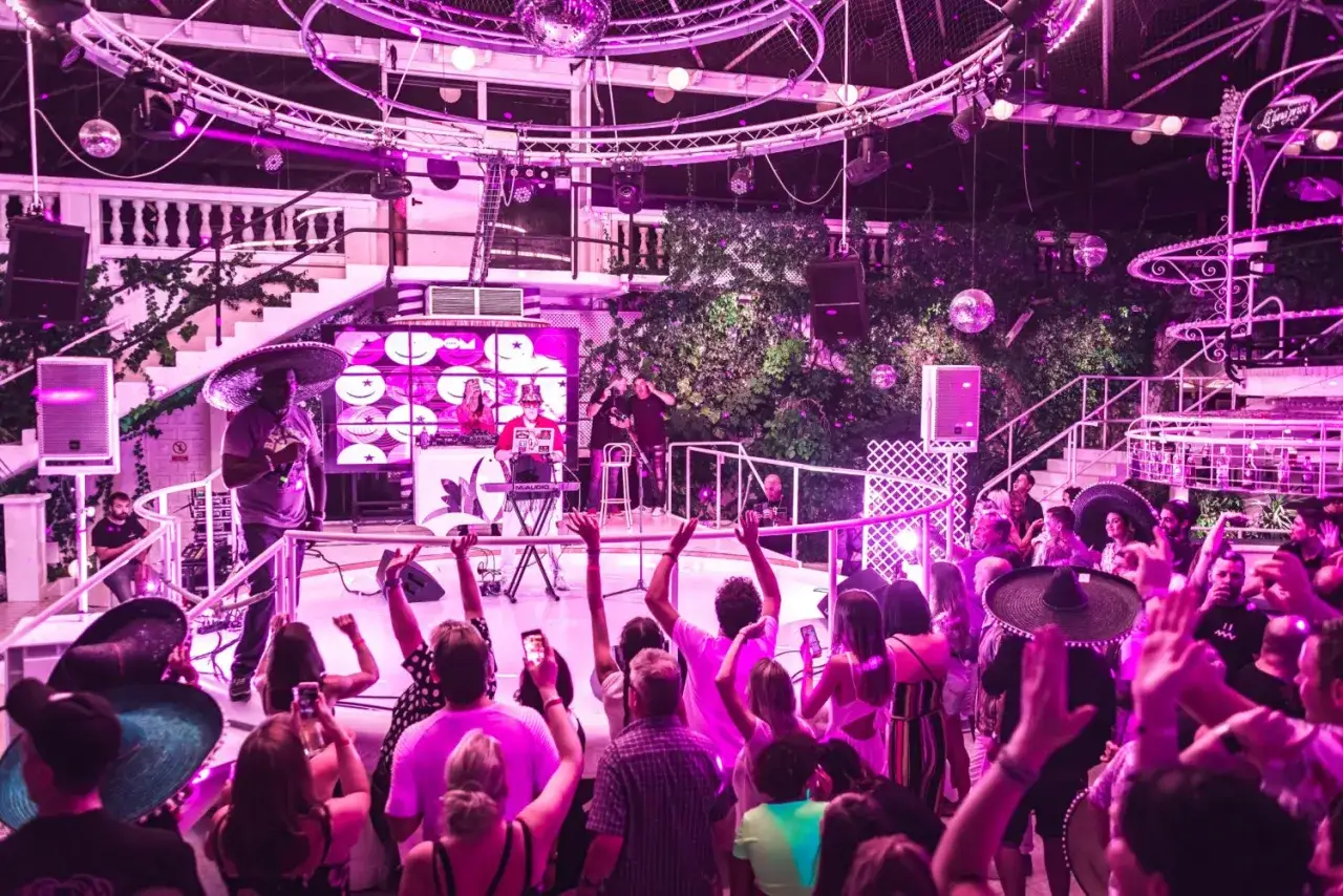 Un concerto in una delle discoteche di Ibiza negli anni '90, con DJ, musicisti e un pubblico festante sotto luci viola e sfere da discoteca.