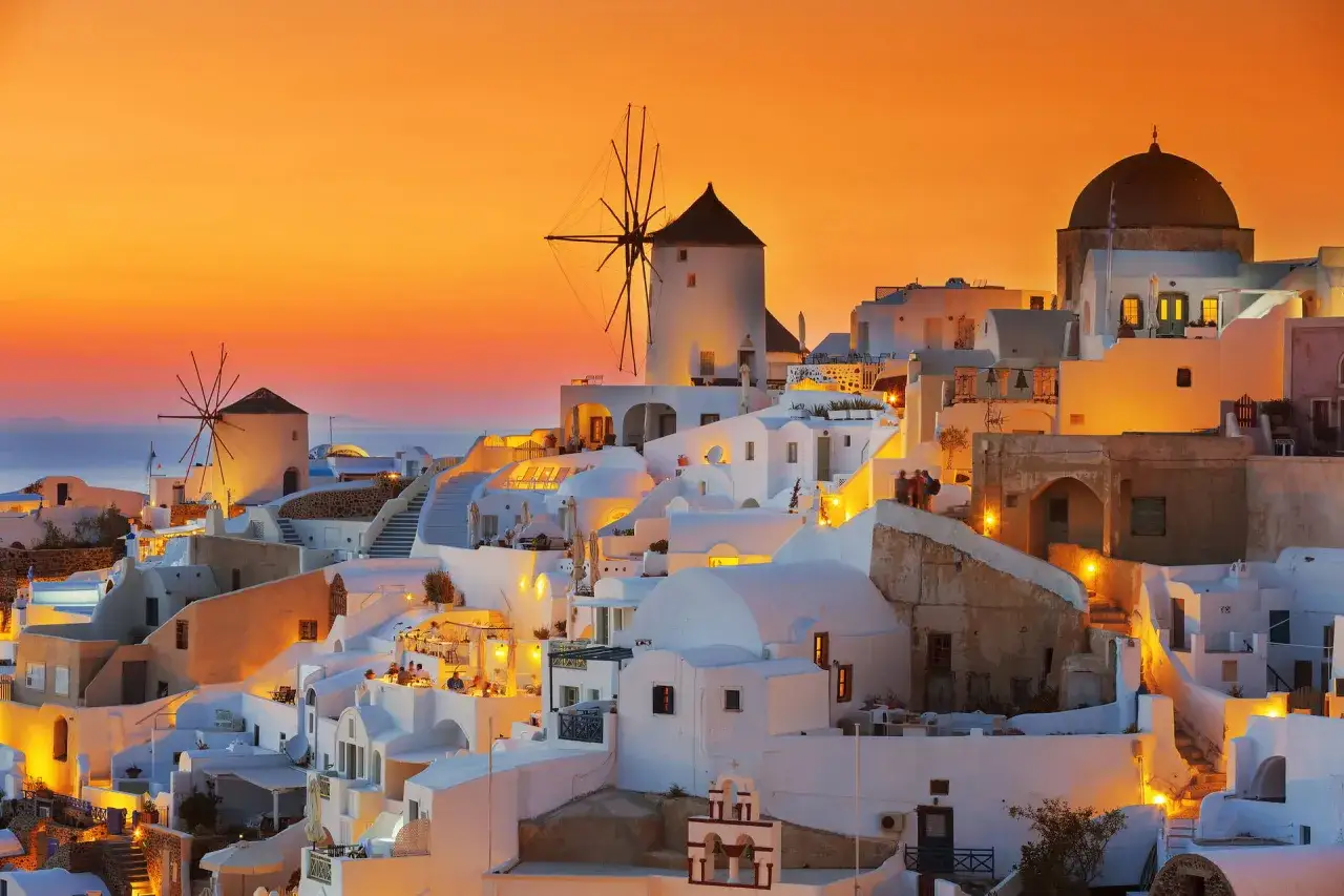 Zachód słońca nad Oia na Santorini. Białe domy, wiatraki i kopuły kościołów oświetlone ciepłym blaskiem. Idealne miejsce na niezapomniane **Santorini wycieczki**.