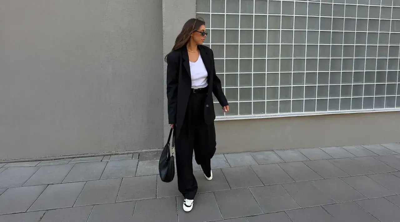 Une femme porte un élégant **outfit pull sans manche** noir, un blazer oversize et un sac à main. Elle marche sur un trottoir devant un mur gris.