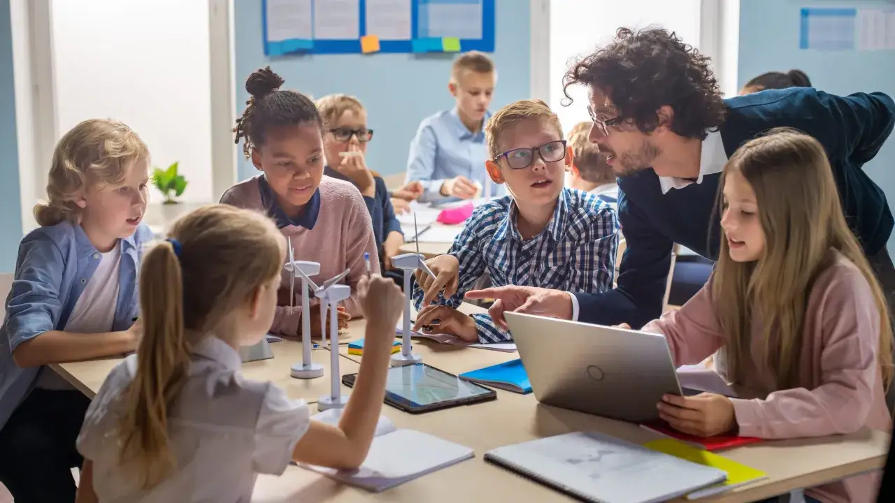 Lehrer erklärt Schülern im **flipped classroom**-Stil Windkraftmodelle. Kinder arbeiten an Tablets und Laptops.