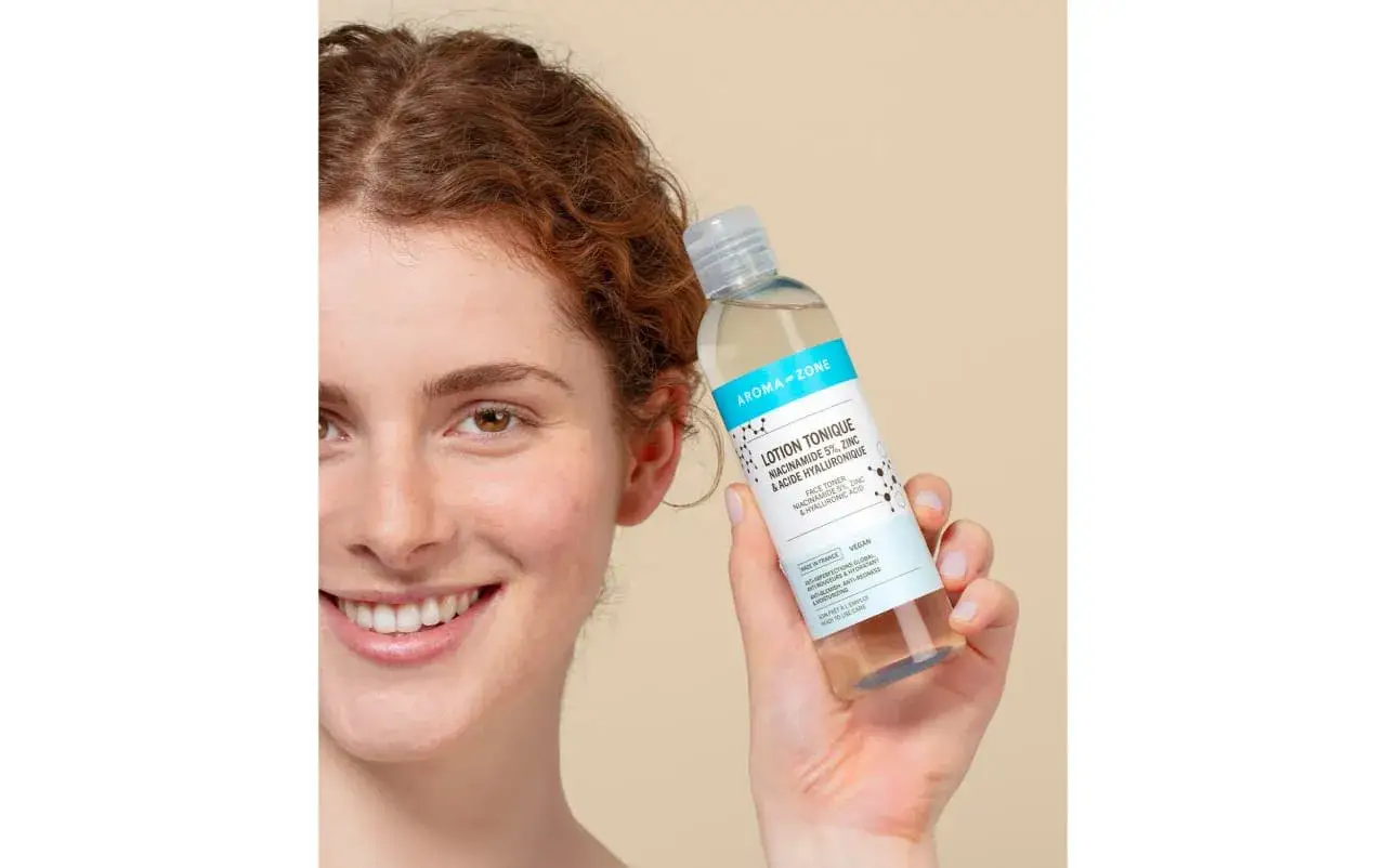 Une femme souriante tient une lotion tonique Aroma-Zone. Ce produit sert &agrave; unifier le teint, r&eacute;duire les imperfections et hydrater.
