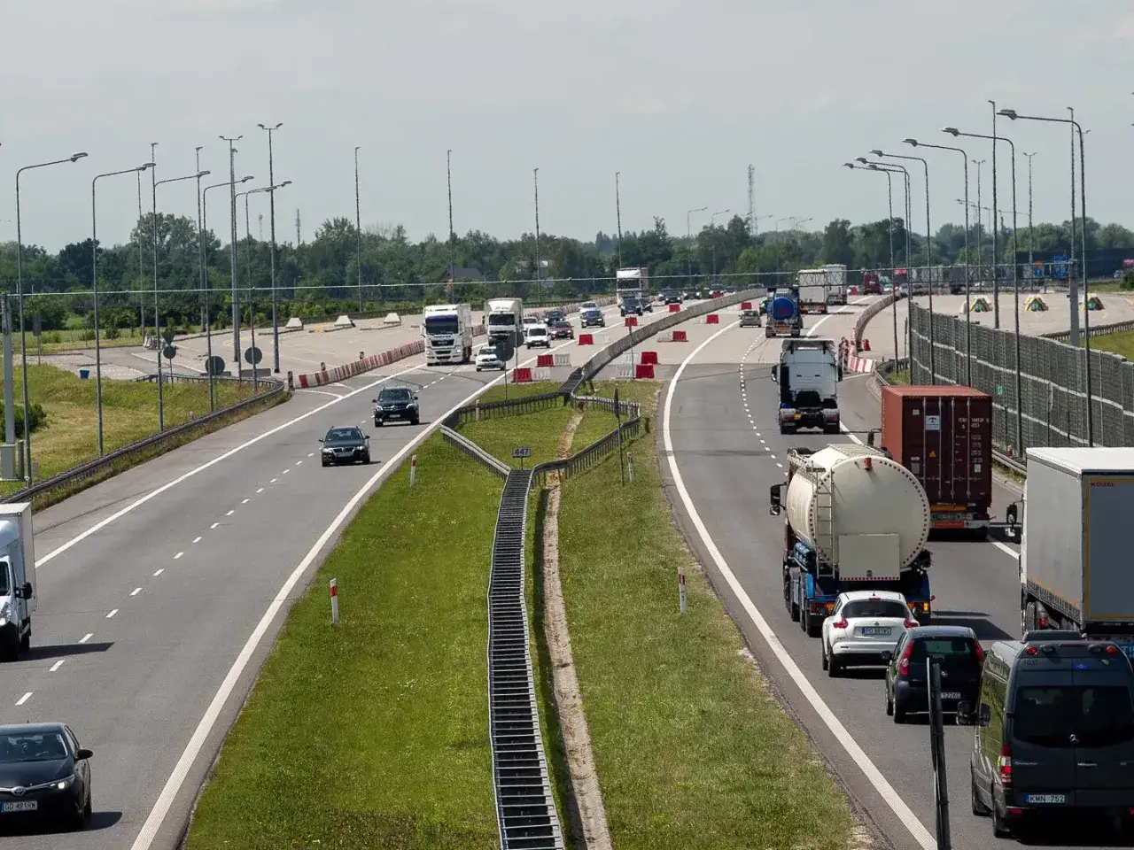 Autostrada z ruchem lewostronnym, pełna ciężarówek i samochodów osobowych. W tle drzewa i niebo.