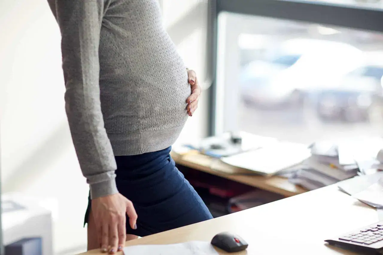 Femme enceinte tenant son ventre, près d'un bureau encombré. Elle pourrait préparer une lettre de démission pendant congé maternité.