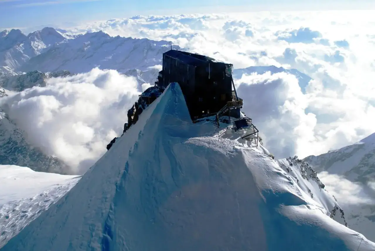 Rifugio più alto d'Europa, un edificio scuro su una vetta innevata, circondato da nuvole e montagne maestose.