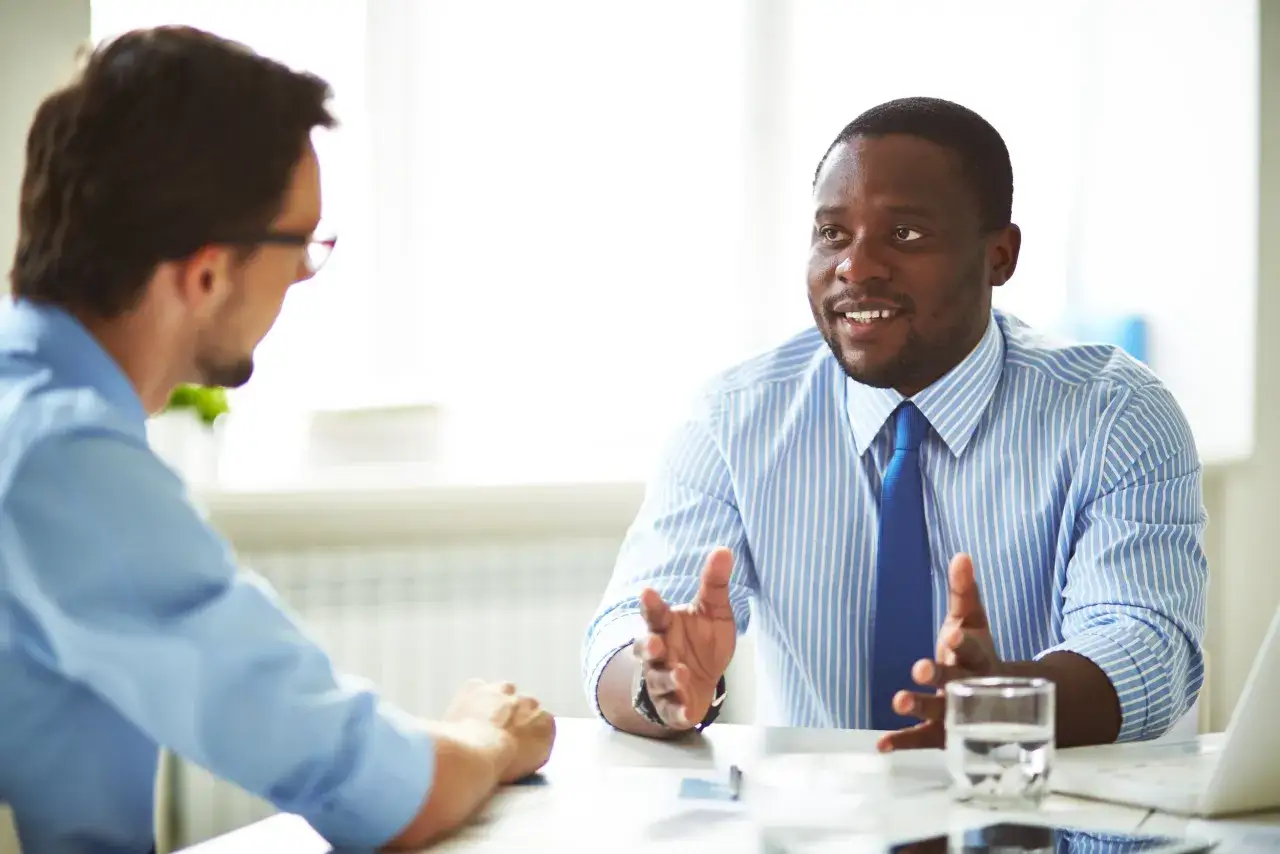 A man in a blue striped shirt and tie gestures while discussing something, embodying effective behavioral leadership in a business meeting.