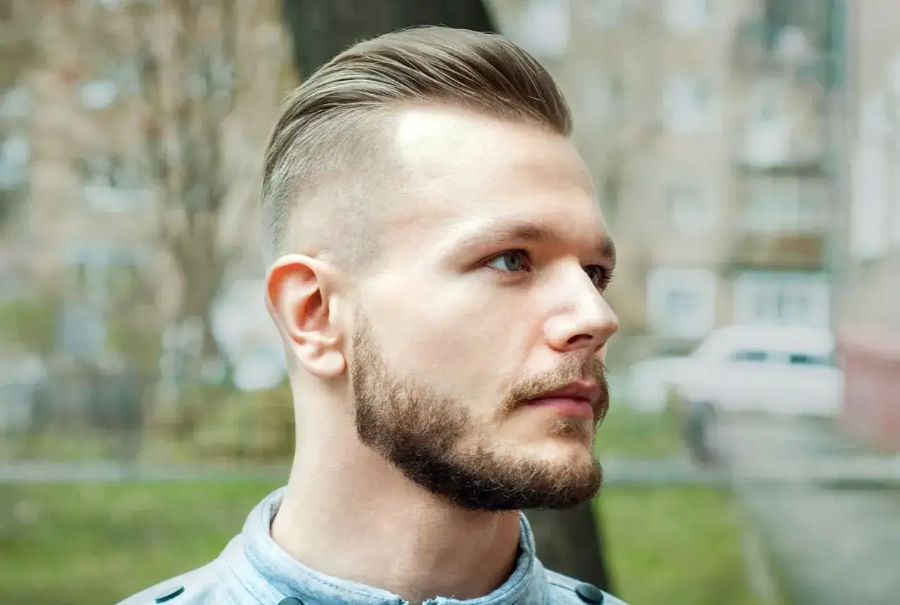 Hombre con pelo liso peinado hacia atrás y barba cuidada, con un corte moderno y afeitado en los laterales.