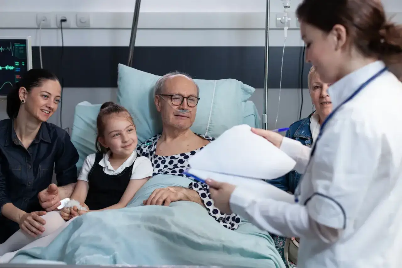 Paciente en cama de hospital con familia. Una auxiliar de enfermer&iacute;a, con su **grado medio de auxiliar de enfermer&iacute;a**, revisa un informe.