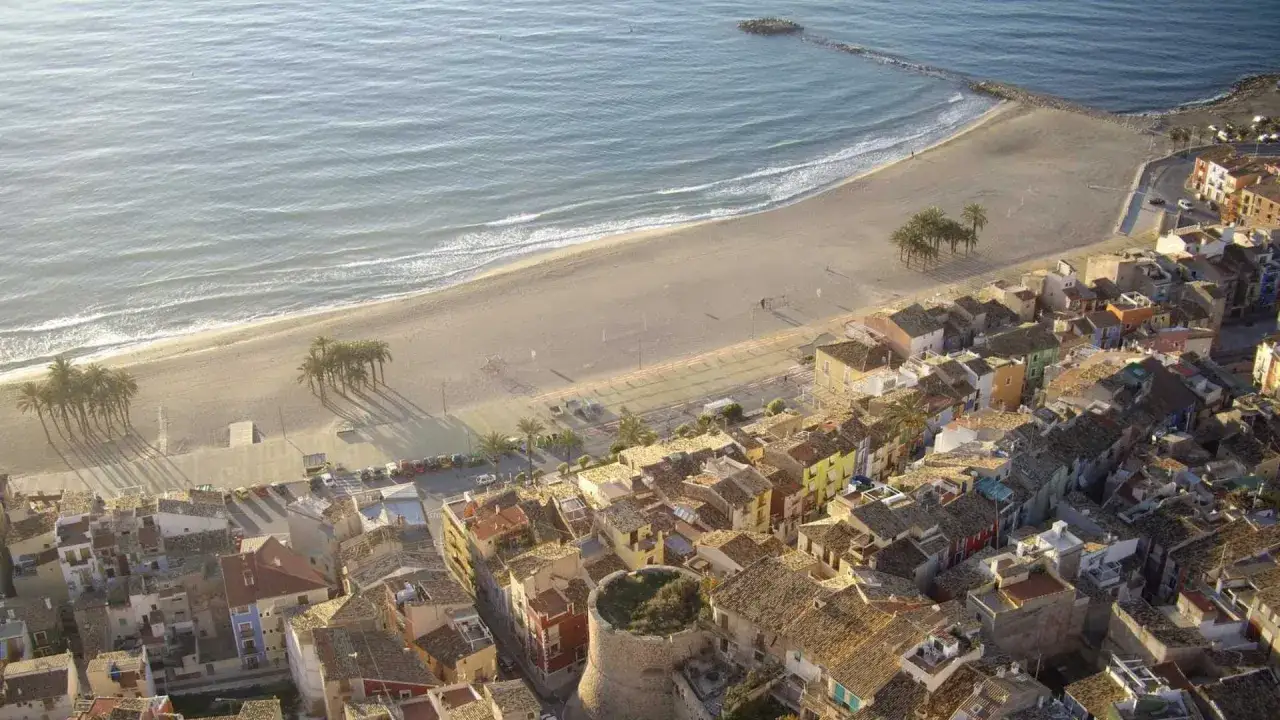 Vista aérea de un pueblo costero con casas coloridas, una playa de arena y el mar. Se ve un antiguo torreón, como si fuera un escenario para el maror fest.