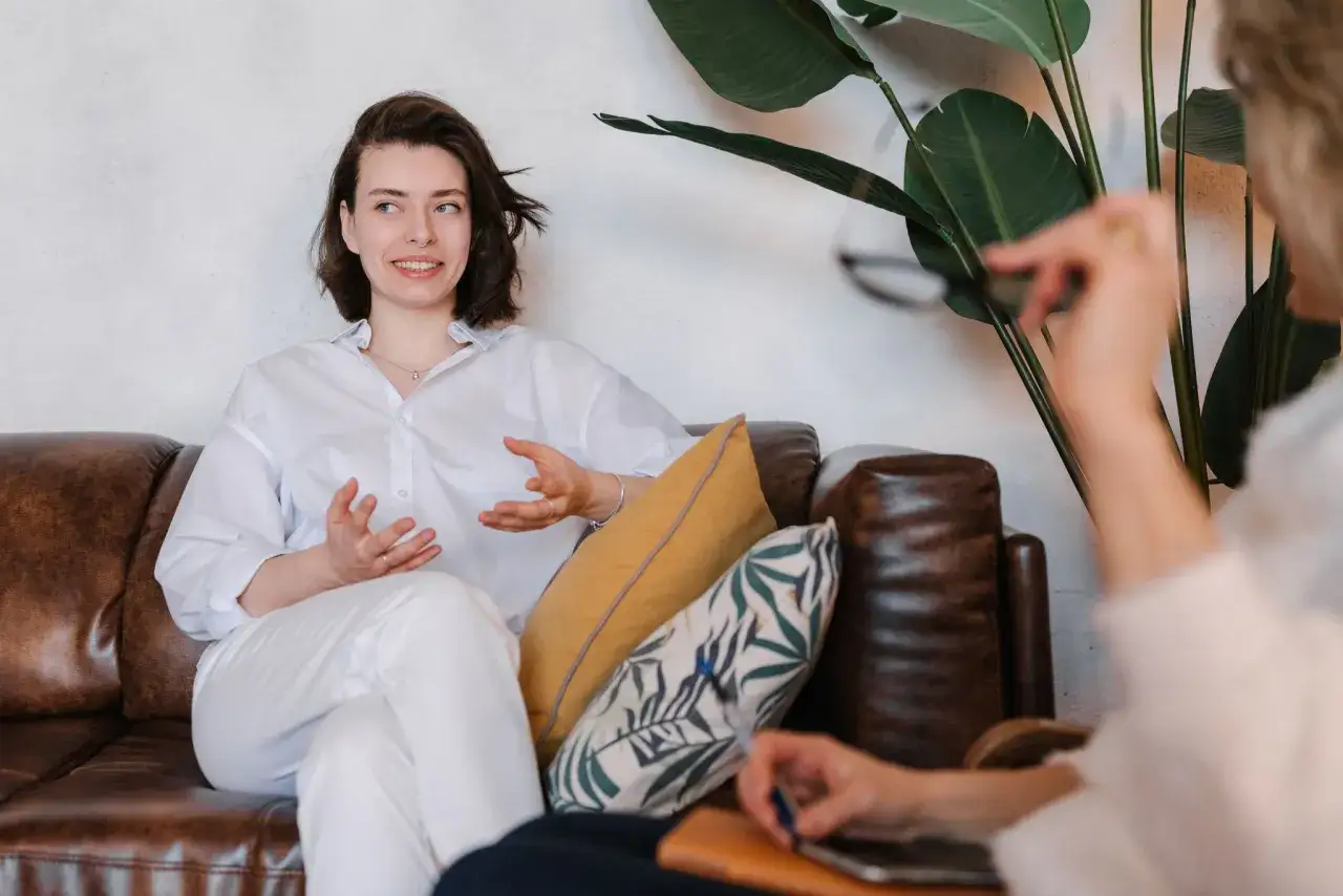 Młoda kobieta z kolczykiem w nosie opowiada o swoich doświadczeniach, siedząc na kanapie. Może to być sesja terapeutyczna lub rozmowa podczas psychologia studia zaoczne.