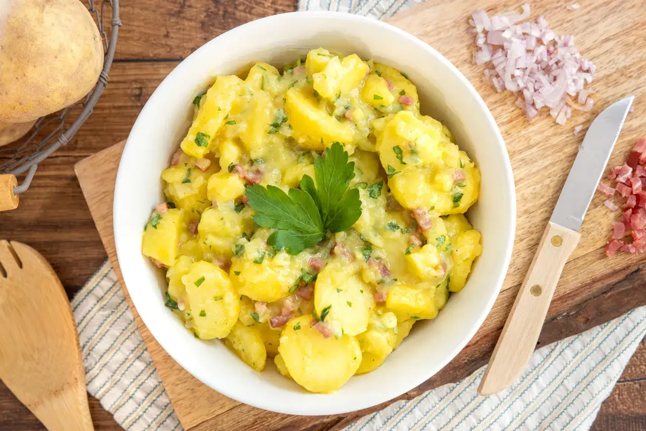 Omas Kartoffelsalat, cremig und mit Speckstücken, garniert mit Petersilie. Daneben liegen Zwiebeln und ein Messer.